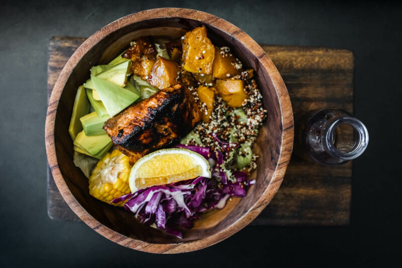 Wooden bowl with fried salmon fish steak, quinoa, avocado, corn, cabbage salad and baked pumpkin. Olive oil in bottle on side. Dark black background, top view. Healthy organic food concept.